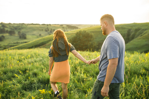 young beautiful couple red-haired girl in a pink dress and green jacket a man in a gray t-shirt and green shorts are having fun in the grass in a field in nature at sunset - Foto, Bild