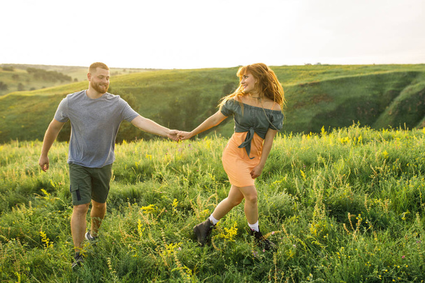 young beautiful couple red-haired girl in a pink dress and green jacket a man in a gray t-shirt and green shorts are having fun in the grass in a field in nature at sunset - Fotografie, Obrázek