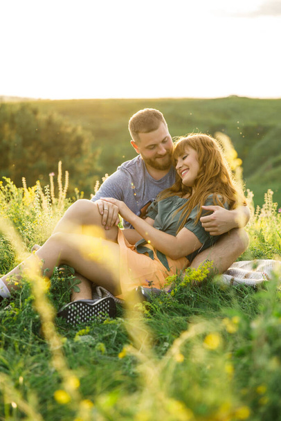 young beautiful couple red-haired girl in a pink dress and green jacket a man in a gray t-shirt and green shorts are having fun in the grass in a field in nature at sunset - Фото, зображення