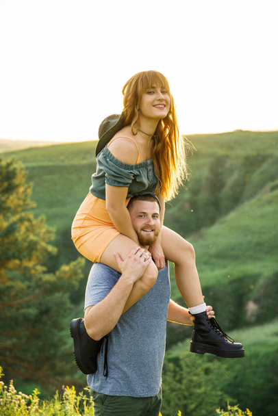 young beautiful couple red-haired girl in a pink dress and green jacket a man in a gray t-shirt and green shorts are having fun in the grass in a field in nature at sunset - Foto, Imagen