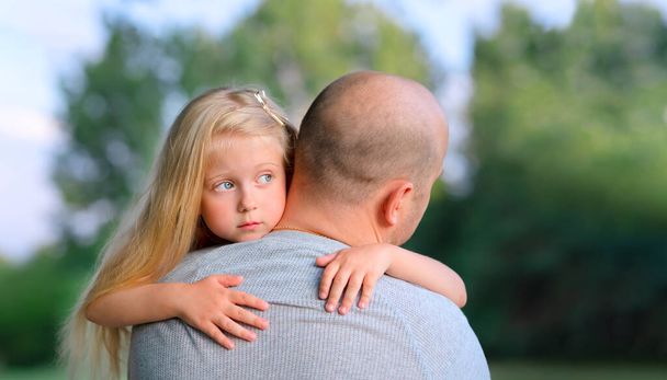 Μικρή ξανθιά λυπημένη κόρη αγκαλιάζει τον μπαμπά του σε εξωτερικό χώρο - Φωτογραφία, εικόνα