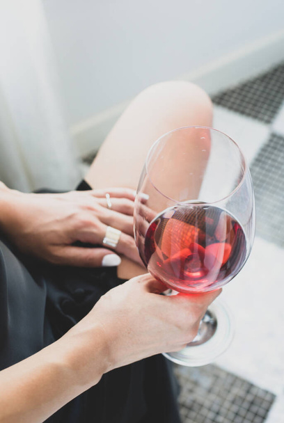 Sklenici červeného vína v ruce dívky v černých koktejlových šatech. Úlomek holých nohou dívky a sklenici vína v její ruce. - Fotografie, Obrázek