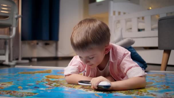 Ein kleiner lächelnder Junge liegt in seinem Zimmer auf dem Boden und erkundet die große Weltkarte durch eine Lupe. Konzept für Reisen, Tourismus und Kindererziehung. Kinder erkunden und entdecken - Filmmaterial, Video