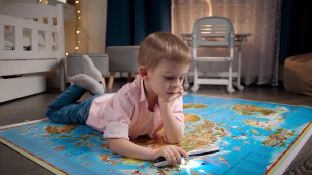 Glimlachend jongetje dat op de vloer ligt en nieuwe landen leert op de grote wereldkaart. Concept van reizen, toerisme en onderwijs voor kinderen. Verkenning en ontdekkingen van kinderen. - Video