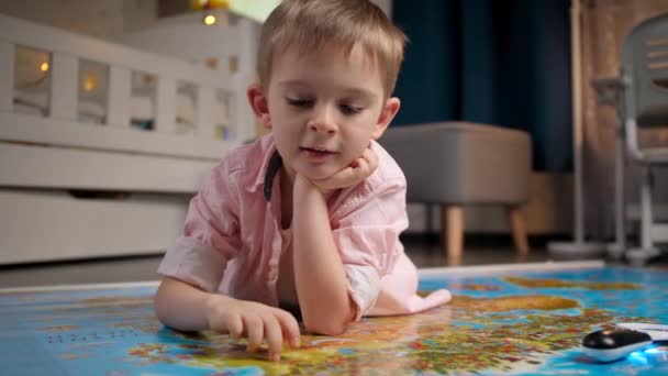 Netter lächelnder Junge, der auf dem Boden liegt und mit den Fingern auf der großen Weltkarte durch Länder geht. Konzept für Reisen, Tourismus und Kindererziehung. Kinder erkunden und entdecken. - Filmmaterial, Video