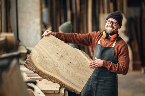 Artisanat barbu adulte positif dans un tablier montrant une grande planche en bois tout en se tenant près de l'établi dans un atelier de menuiserie - Photo, image