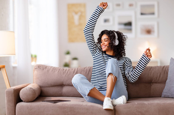 Delighted ethnic female in headphones smiling with closed eyes and dancing while listening to music on couch at home - Foto, Bild