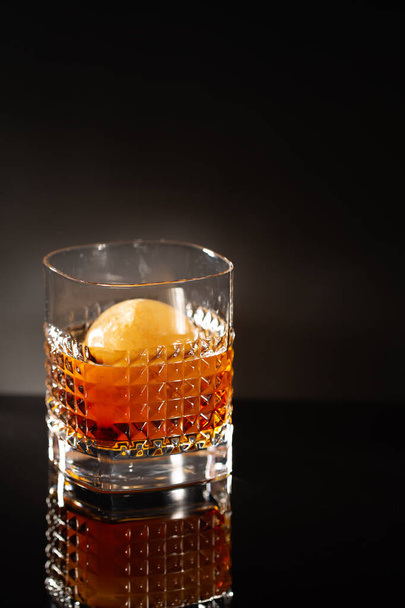 Elegant chrystal glass of Whiskey with whiskey ice ball on black with reflection.Close-up of noble acoholic drink.Minimalistic style. - Fotó, kép