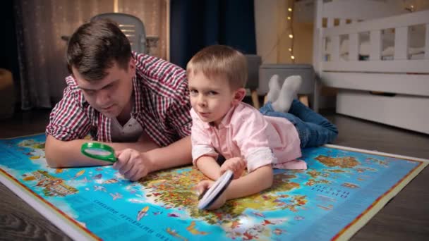Hauska kuva onnellinen msiling isä pieni poika tutkii maailmankarttaa ja leikkii suurennuslasilla. Käsite matkailu, matkailu ja lasten koulutus. Lapset etsintä ja löytö - Materiaali, video