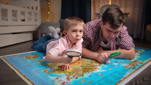 Gelukkige kleine jongen met vader liggend op grote kaart en kijkend door vergrootglas. Concept van reizen, toerisme en onderwijs voor kinderen. Verkenning en ontdekking van kinderen - Video