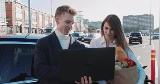 Εστιασμένος άντρας και νέα όμορφη γυναίκα κρατώντας φορητό υπολογιστή στα χέρια. Νέοι επαγγελματίες με τη βοήθεια υπολογιστή στο πάρκινγκ κοντά στο σούπερ μάρκετ ψωνίζουν. Σταθμός φόρτισης αυτοκινήτων - Πλάνα, βίντεο