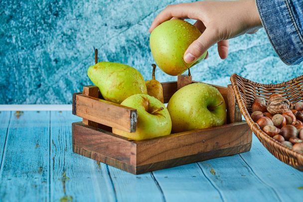Apples and pear in wooden basket and bowl of hazelnuts on blue background - Foto, Bild