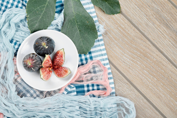 Talíř z celých a nakrájených černých fíků, list a modré a růžové ubrusy na dřevěném stole - Fotografie, Obrázek