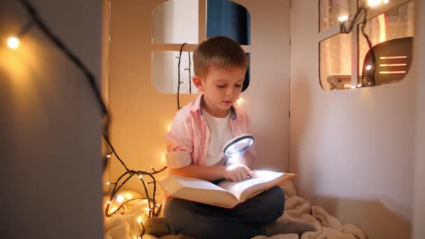 Lindo niño leyendo libro con antorcha en su casa de cartón de juguete por la noche. Concepto de educación y lectura infantil en cuarto oscuro - Metraje, vídeo