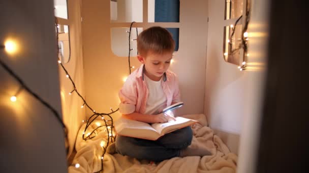 Cute little boy sitting in his toy house and reading book with magnifying glass and torch. Concept of child education and reading in dark room - Footage, Video
