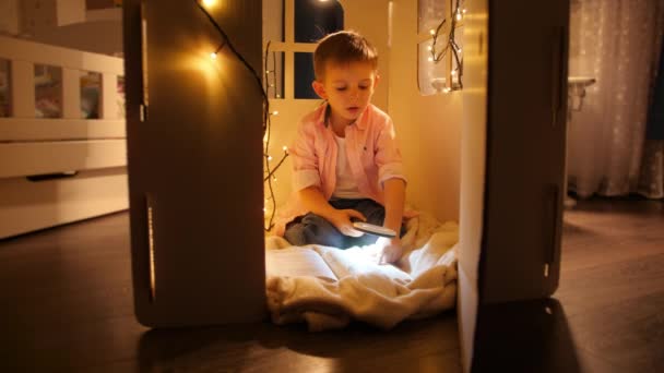 Dolly shot van kleine jongen met behulp van zaklamp en vergrootglas om boek te lezen 's nachts. Concept van kinderopvoeding en lezen in de donkere kamer - Video