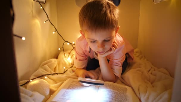 Portrait of little smart boy reading big story book at night. Child playing in toy cardboard house. Concept of child education and reading in dark room - Footage, Video