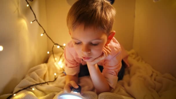 Dolly shot van schattige jongen lezen boek tijdens het spelen in zijn speelgoed huis 's nachts. Concept van kinderopvoeding en lezen in de donkere kamer - Video