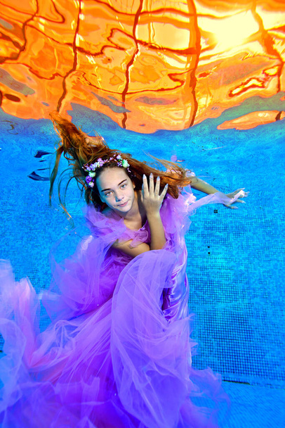 A young teenage girl has fun and swims in the pool in a beautiful purple dress with her hair down and a flower wreath on her head, and looks at the camera. Fashion portrait. Concept. - 写真・画像