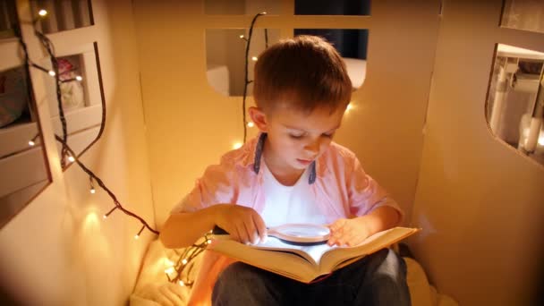 Portret van een jongetje met een kijker en vergrootglas terwijl hij 's nachts het boek leest. Concept van kinderopvoeding en lezen in de donkere kamer - Video