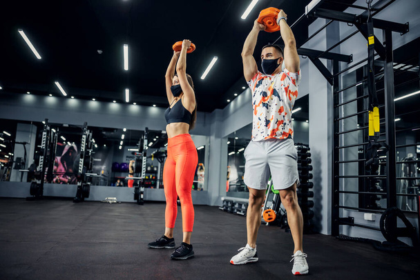 Sporty couple in shape with face masks standing in gym and lifting weights. Healthy lifestyle, sport during corona, bodybuilding - Photo, Image