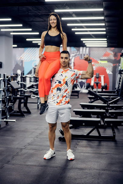Starker muskulöser Sportler, der Frau auf Schulter hält und in einem Fitnessstudio posiert. Bodybuilding, gesundes Leben - Foto, Bild