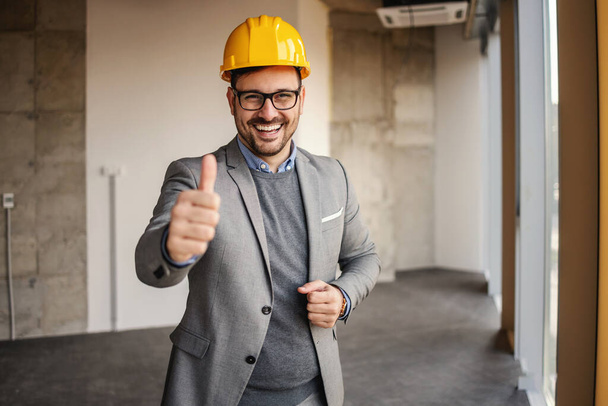 Arquiteto sorridente em pé no edifício em processo de construção e mostrando polegares para cima. - Foto, Imagem