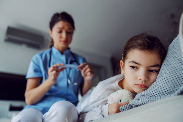 Krankes kleines Mädchen, das zu Hause auf dem Bett liegt. Im verschwommenen Hintergrund blickt eine Krankenschwester auf das Thermometer. - Foto, Bild