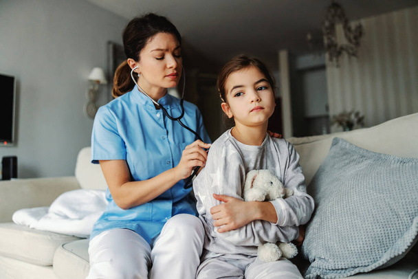 Médico sentado no sofá ao lado da menina e examinando seus pulmões com estetoscópio. Médico em serviço em casa. - Foto, Imagem