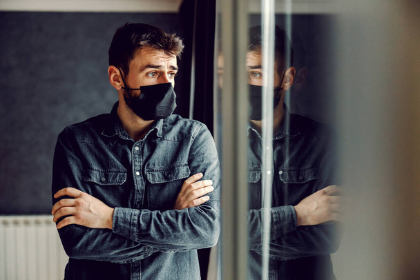 Испуганный человек в маске, стоящий у окна с скрещенными руками во время блокировки коронного вируса. - Фото, изображение