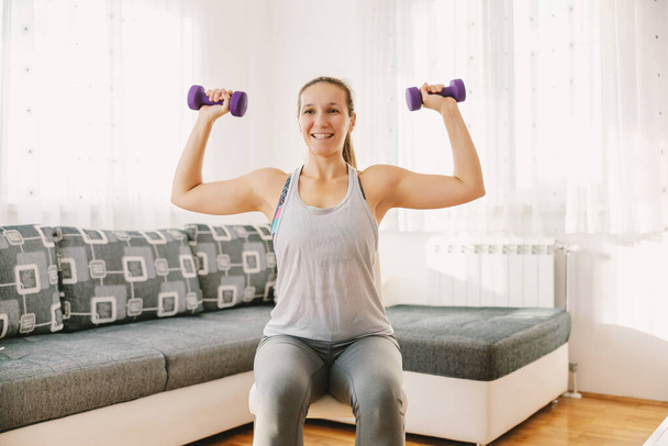 Smiling sportswoman sitting on the chair in her apartment and doing exercises for biceps. If you can't go to the gym you can make your own at home. - Fotoğraf, Görsel