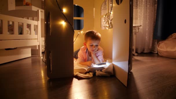 Happy smiling boy lying on floor at hos toy house or tent and reading with flashlight at night. Concept of child education and reading in dark room - Footage, Video