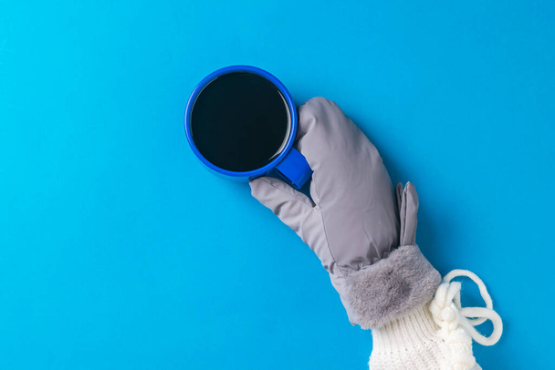 Eine Hand in einem warmen Handschuh mit einer Tasse heißen Kaffees auf blauem Hintergrund. Heißgetränk und Handschuhe. - Foto, Bild