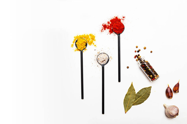 Various spices in black spoons. Salt, curry, turmeric, ground paprika, bay leaf, garlic, peppercorns flat lay on a white background - Photo, Image