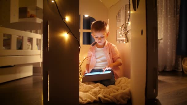 Little smart boy sitting in toy cardboard house and using tablet computer. Concept of child education and studying at night - Footage, Video