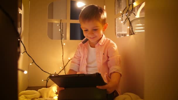 Muotokuva onnellinen msiling poika käyttää tablet-tietokone ja selailu internetissä pelatessaan hänen lelu pahvi talon tai teltan yöllä. Lastenkasvatuksen ja opiskelun käsite yöllä - Materiaali, video