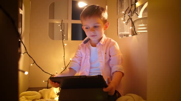 Pienen pojan muotokuva katsomassa piirrettyjä tabletin tietokoneella yöllä, kun hän istuu pienessä lelutalossa. Lastenkasvatuksen ja opiskelun käsite yöllä - Materiaali, video