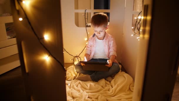 Dolly disparo de niño sonriente sentado en su pequeña casa de juguetes y el uso de la tableta de ordenador. Concepto de educación infantil y estudio nocturno - Imágenes, Vídeo