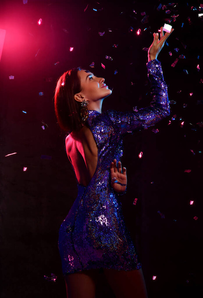 Sexy brunette woman in glittering backless tight mini dress and stands sideways to camera under confetti fall touching - Fotografie, Obrázek