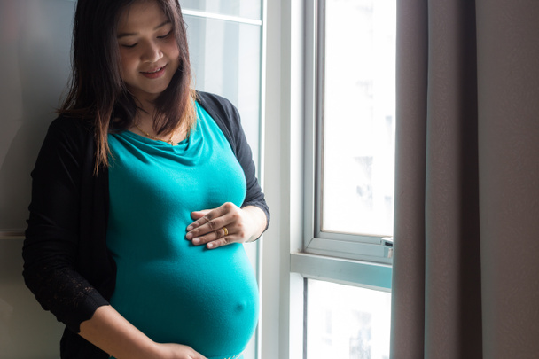 Zwangere vrouwen houden buik staan in de buurt van gordijn in de kamer - Foto, afbeelding