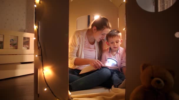 テントやおもちゃの段ボールの家に座って、夜に懐中電灯で本を読んでいる若い母親と幸せな笑顔の男の子。夜間に一緒に時間を持つ子供の教育と家族の概念 - 映像、動画