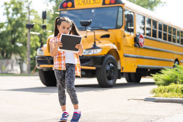 imádnivaló kis iskolás lány az iskolabusz mellett és a kamerába néz. - Fotó, kép