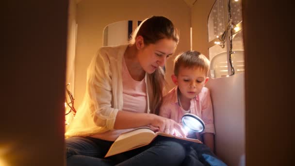Portrait of smiling young mother with little boy reading book at night while playing in toy house. Concept of child education and family having time together at night - Footage, Video
