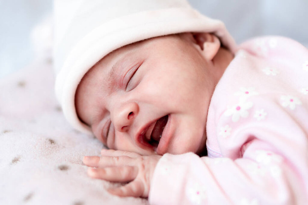 Childhood, illness, colic, bloating, motherhood, health concepts - Close up restless, worried sad newborn baby girl in hat sleeps crying scream have stomachache laying on tummy on bed with open mouth - Foto, immagini