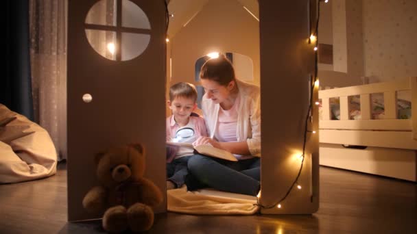 Dolly tiro de jovem mãe lendo livro para seu filho enquanto sentado em pequena casa de brinquedo. Conceito de educação infantil e família tendo tempo juntos à noite - Filmagem, Vídeo