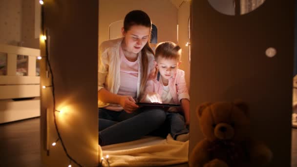 テントやおもちゃの家で遊んでいる若い母親と小さな笑顔の男の子とタブレットコンピュータを使用しています。夜間に一緒に時間を持つ子供の教育と家族の概念 - 映像、動画