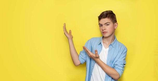 The boy was a teenager in a blue shirt and white T-shirt raised his hands and pointed to a free space for text on a yellow background. - Fotoğraf, Görsel