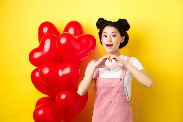 Glückliches asiatisches Mädchen zeigt Herzzeichen in der Nähe romantischer Luftballons, wartet auf die wahre Liebe am Valentinstag, blickt hoffnungsvoll in die Kamera, gelber Hintergrund - Foto, Bild