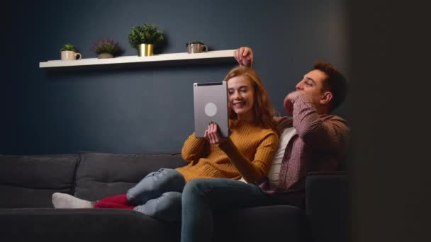 Pareja joven tumbada en un sofá relajándose y abrazándose mientras ve algo en una tableta digital - Imágenes, Vídeo