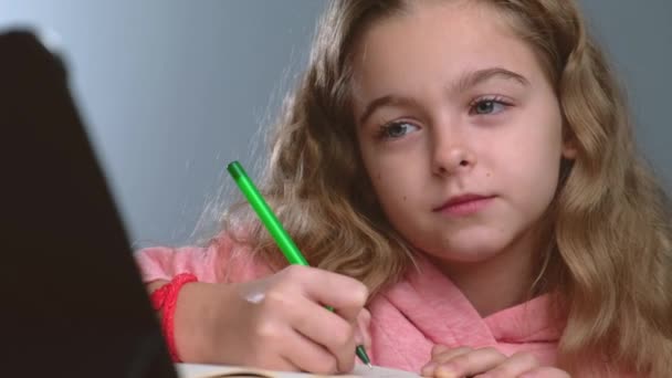 Primer plano video de una chica rubia caucásica teniendo lecciones en línea usando una tableta y escribiendo el tema en el copybook - Metraje, vídeo